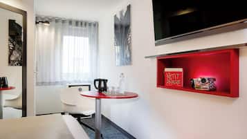 Una habitación de hotel con una cama, una mesita con hervidor, una silla, un televisor y un estante rojo con libros y una cámara.