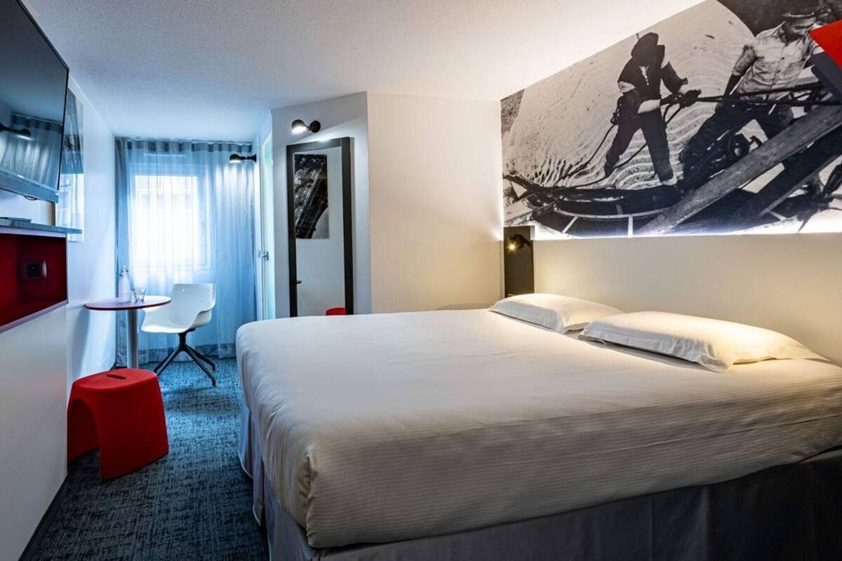 Et hotelværelse med seng, skrivebord, stol, fjernsyn og et stort billede på væggen.