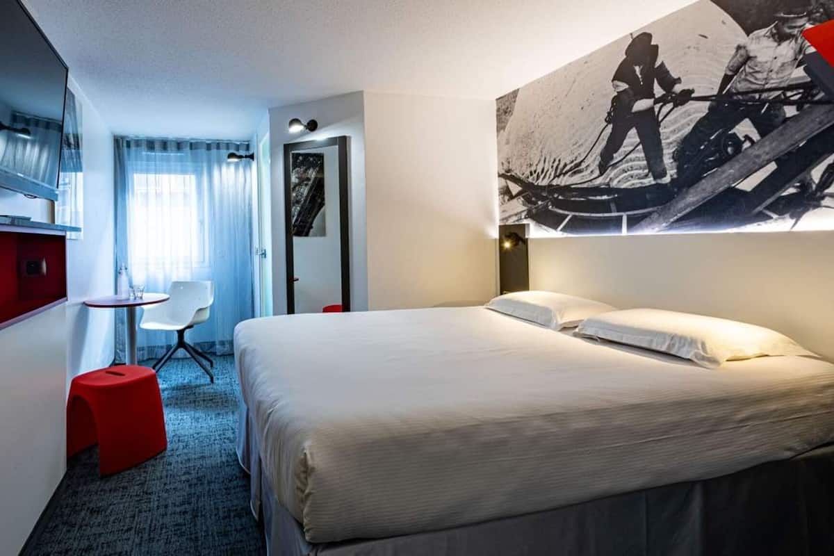 Ett hotellrum med en säng, ett skrivbord, en stol, en tv och en stor affisch på väggen.