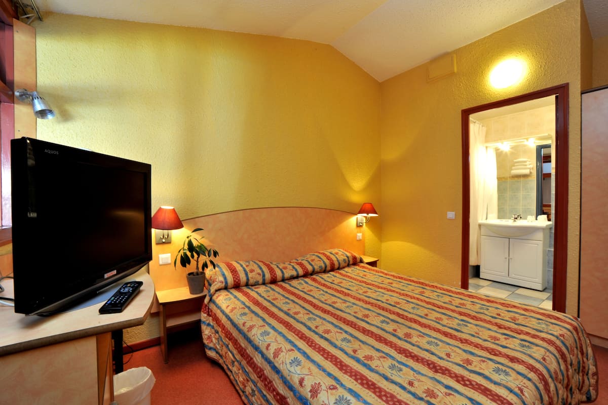 Quarto de hotel com cama, televisão, escrivaninha e banheiro visível por uma porta aberta.