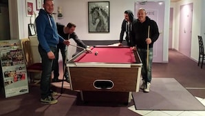 Une table de billard dans une pièce où l’on aperçoit, au mur, une œuvre d’art encadrée, une porte au fond et un présentoir à magazines.