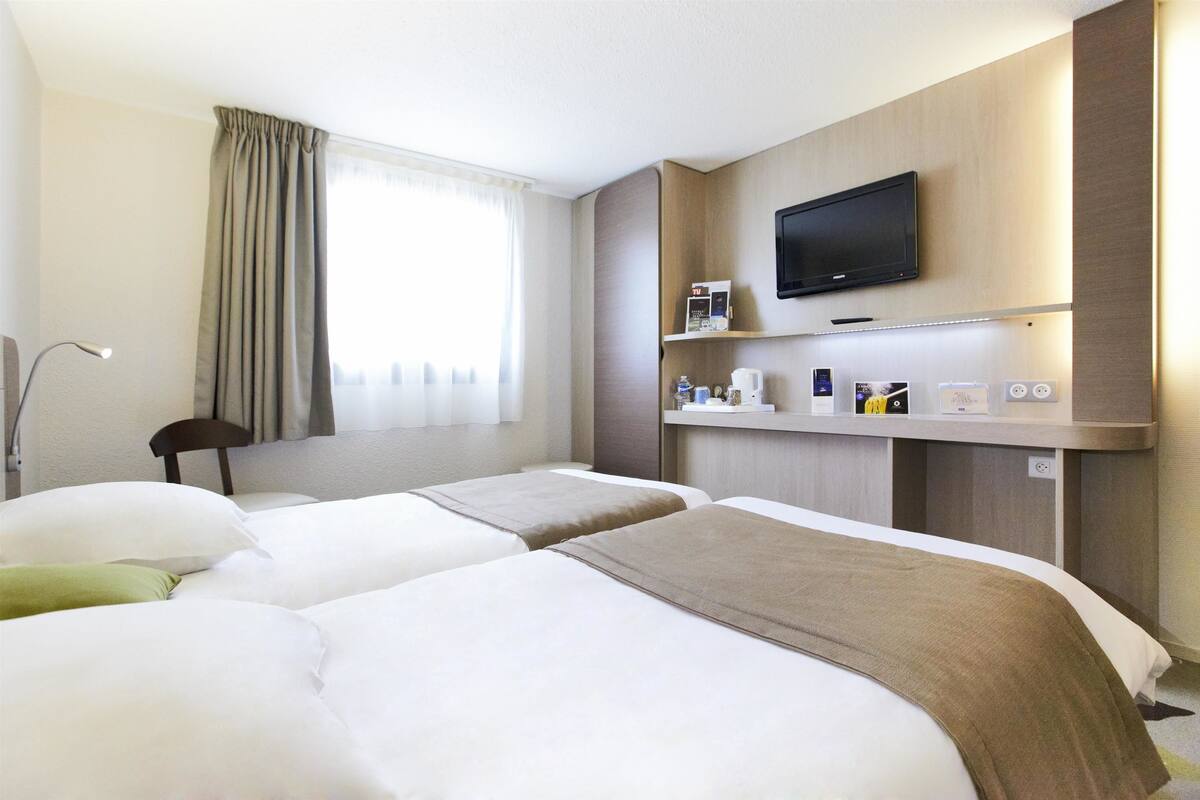 A hotel room with two beds, a TV mounted on the wall, a bedside table with a coffee maker, and a window with curtains.