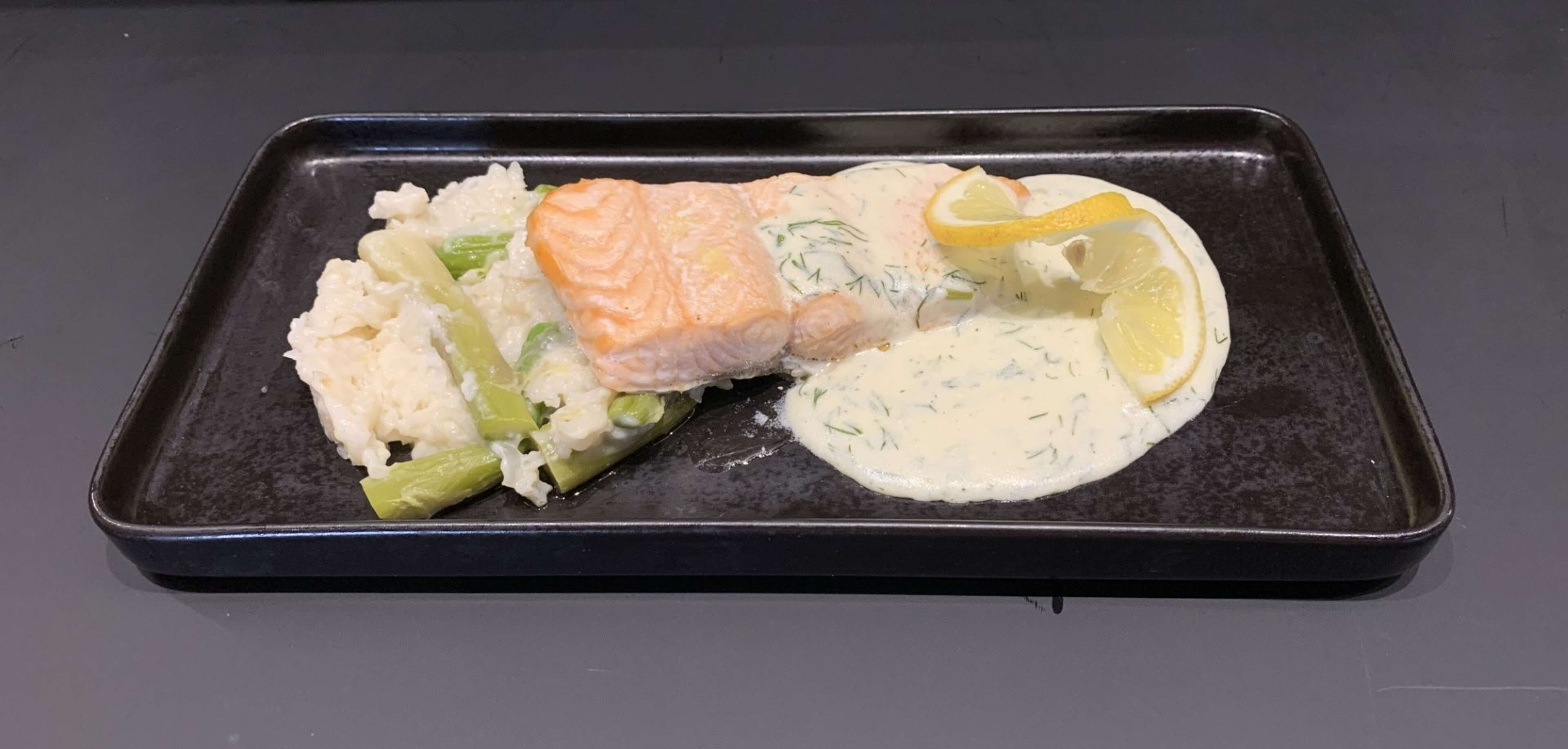 Una bandeja negra rectangular con un filete de salmón, verduras verdes y un gajo de limón sobre una superficie oscura.