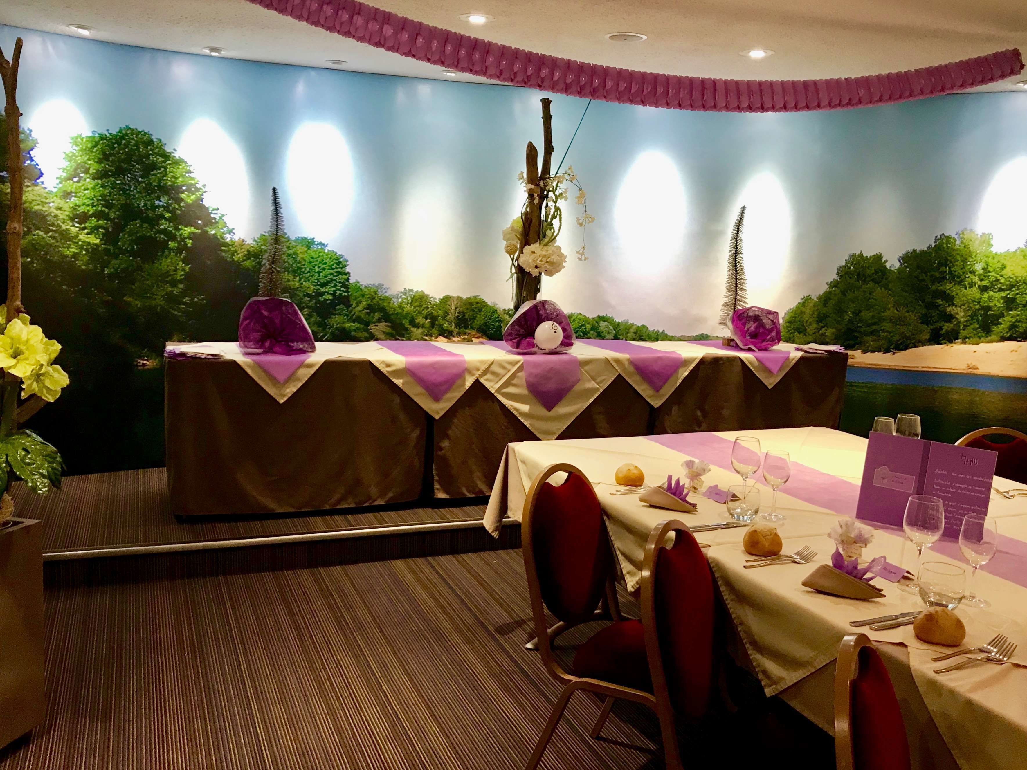 A restaurant with a view of a painted landscape, tables set with white tablecloths and purple napkins, and red chairs.