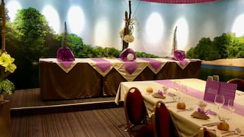 Un restaurante con vistas a un paisaje pintado, mesas dispuestas con manteles blancos y servilletas moradas, y sillas rojas.