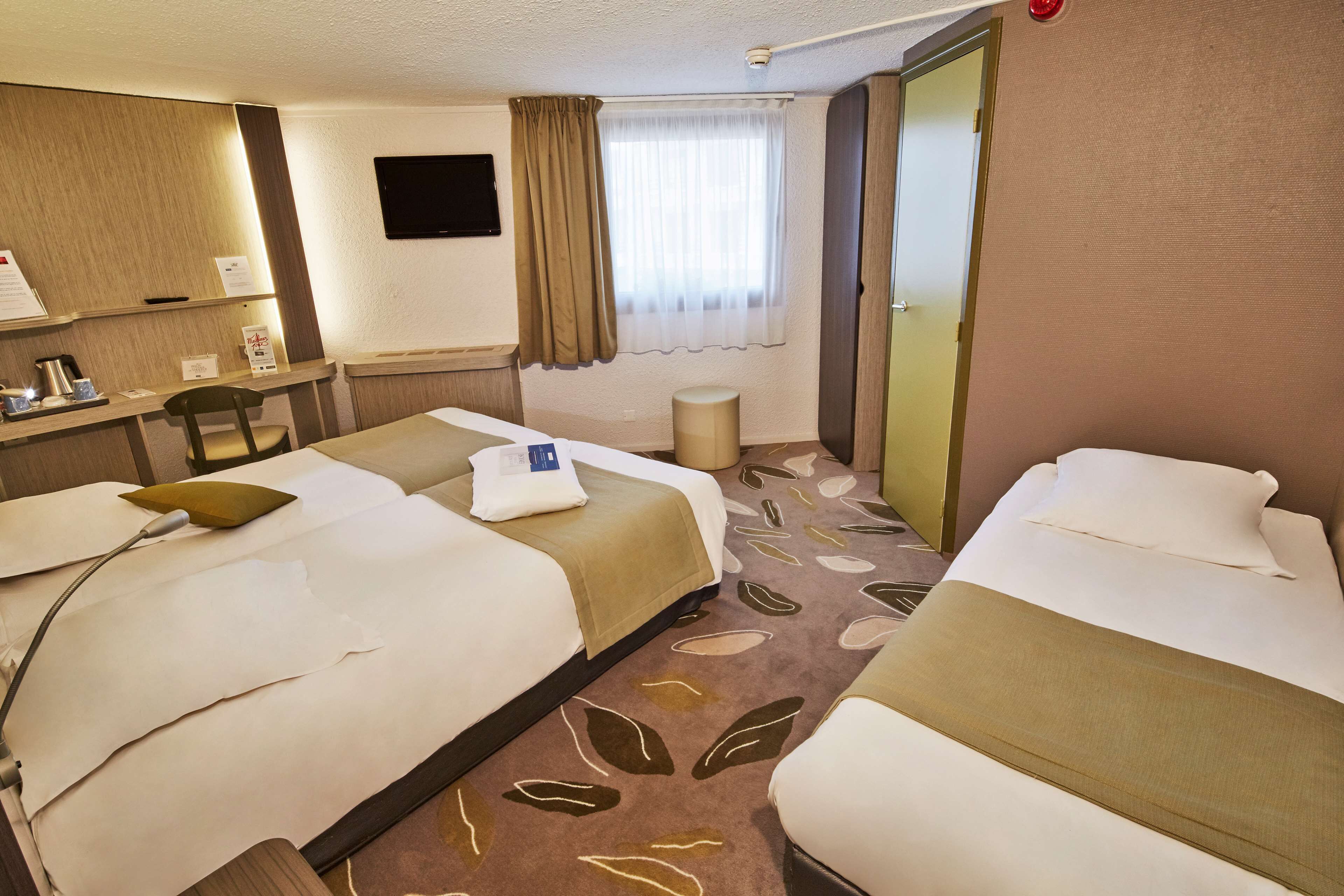 Habitación de hotel con dos camas, un escritorio con una silla, un televisor de pantalla plana, una ventana con cortinas y una alfombra estampada con hojas.
