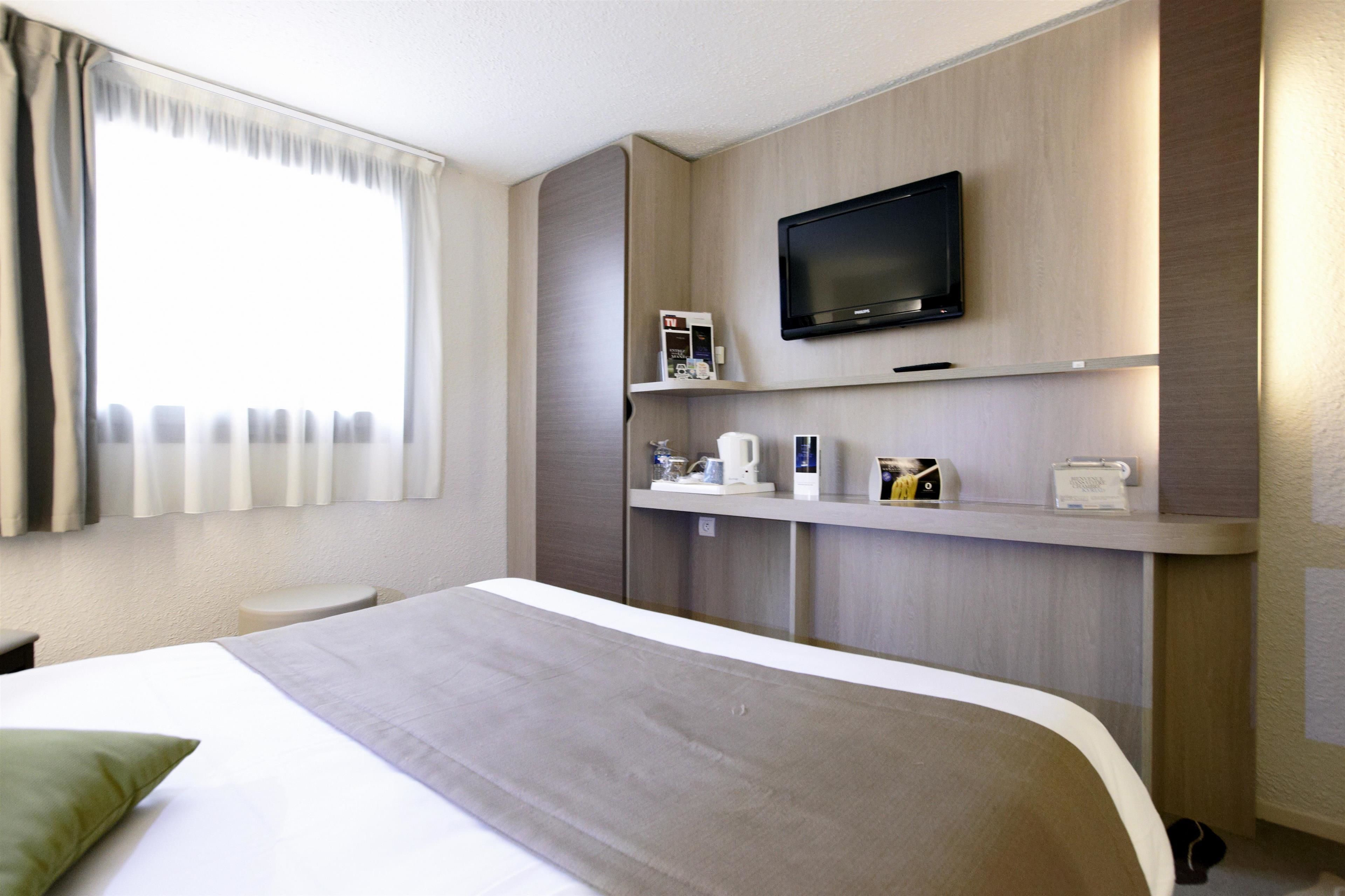 A hotel room with a bed, a television mounted on the wall, a small table, and a window with curtains.