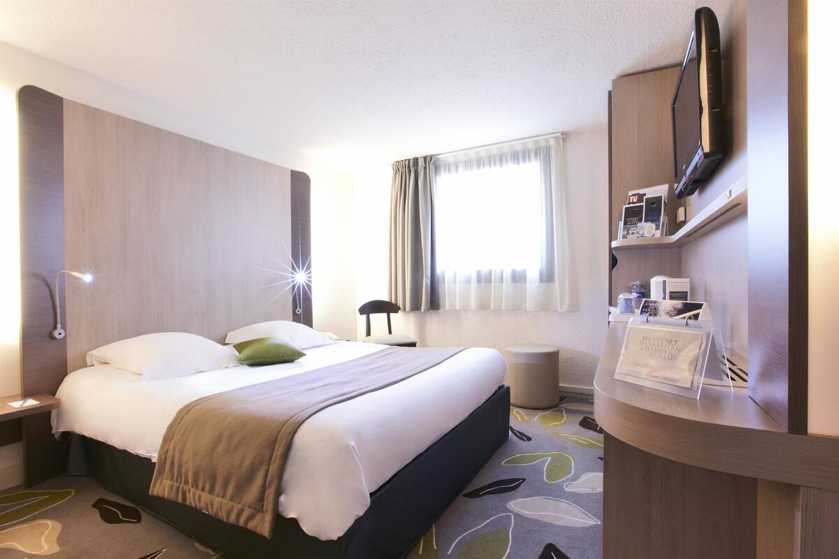 A hotel room with a bed, bedside table, lamp, TV, and a reception desk with a brochure.