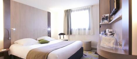 A hotel room with a bed, bedside table, lamp, TV, and a reception desk with a brochure.