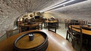 Un interior con paredes de piedra, mesas y sillas redondas, un barril de madera y un televisor de pantalla plana.
