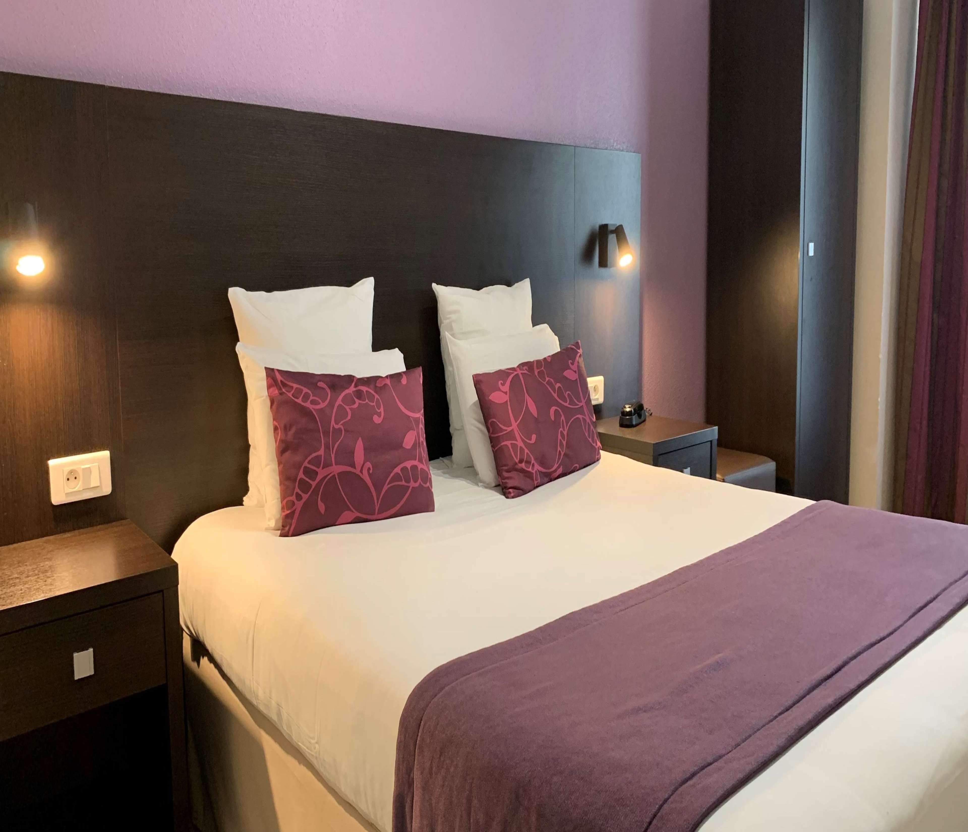 A hotel room with a bed, two bedside tables, a nightstand, and a wall-mounted lamp.