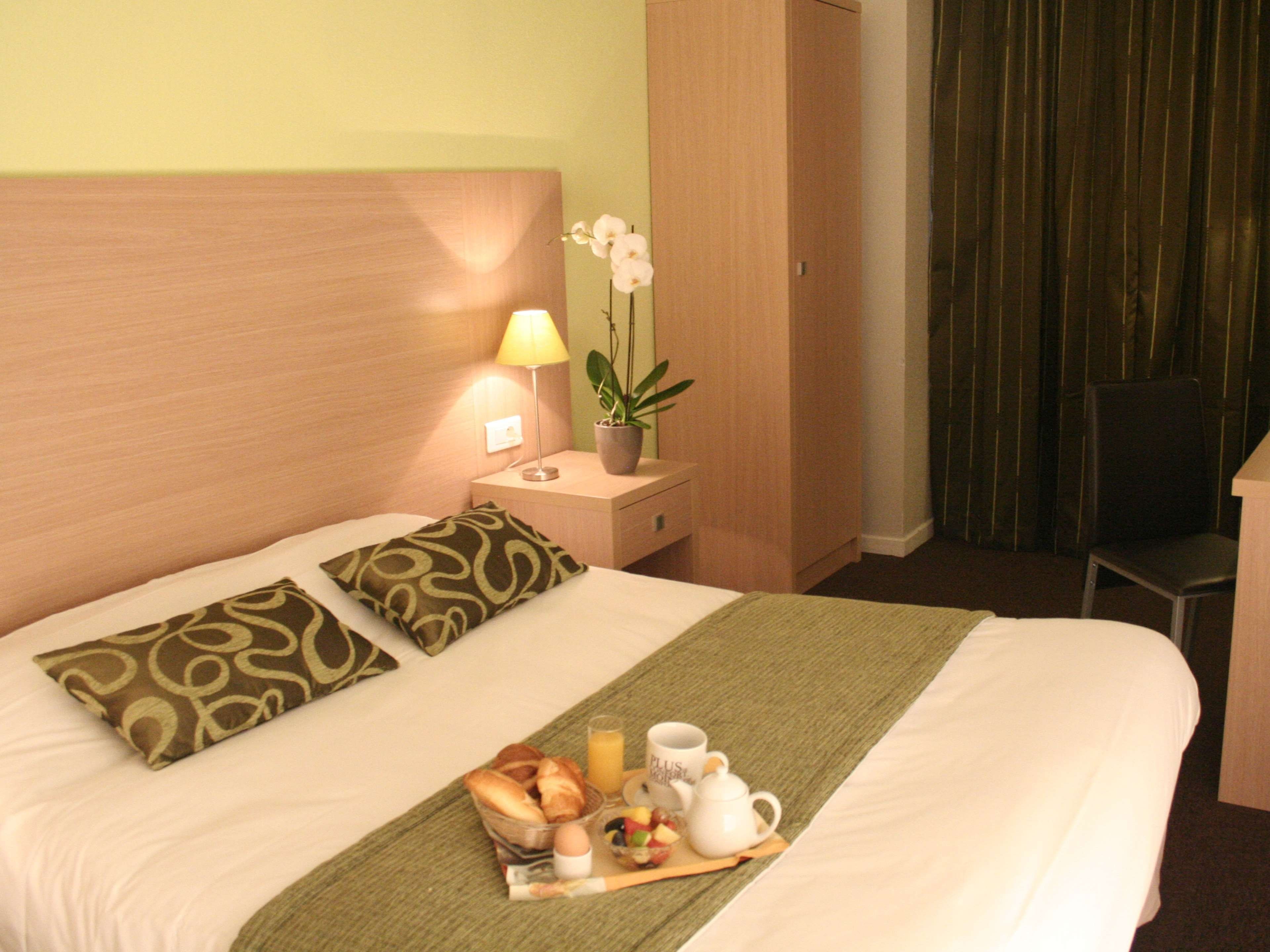 A hotel room with a bed, a nightstand with a lamp, a potted plant, and a breakfast tray.