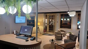 Un mostrador de recepción con una computadora, un teléfono y un teclado. Hay sillas y una peque ña mesa con una planta. Se ve una ventana grande con un letrero de bienvenida.