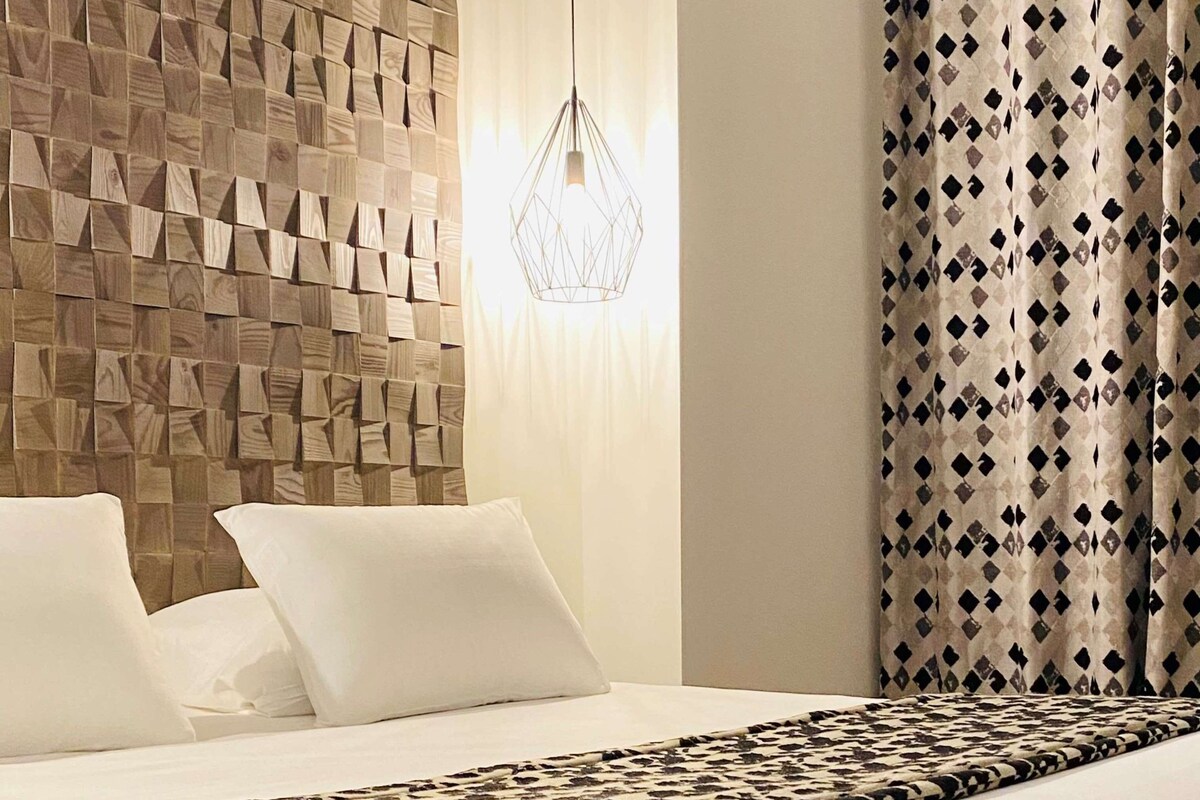 A hotel room with a wooden textured wall, a geometric pendant light, a patterned bedspread, and a patterned curtain.