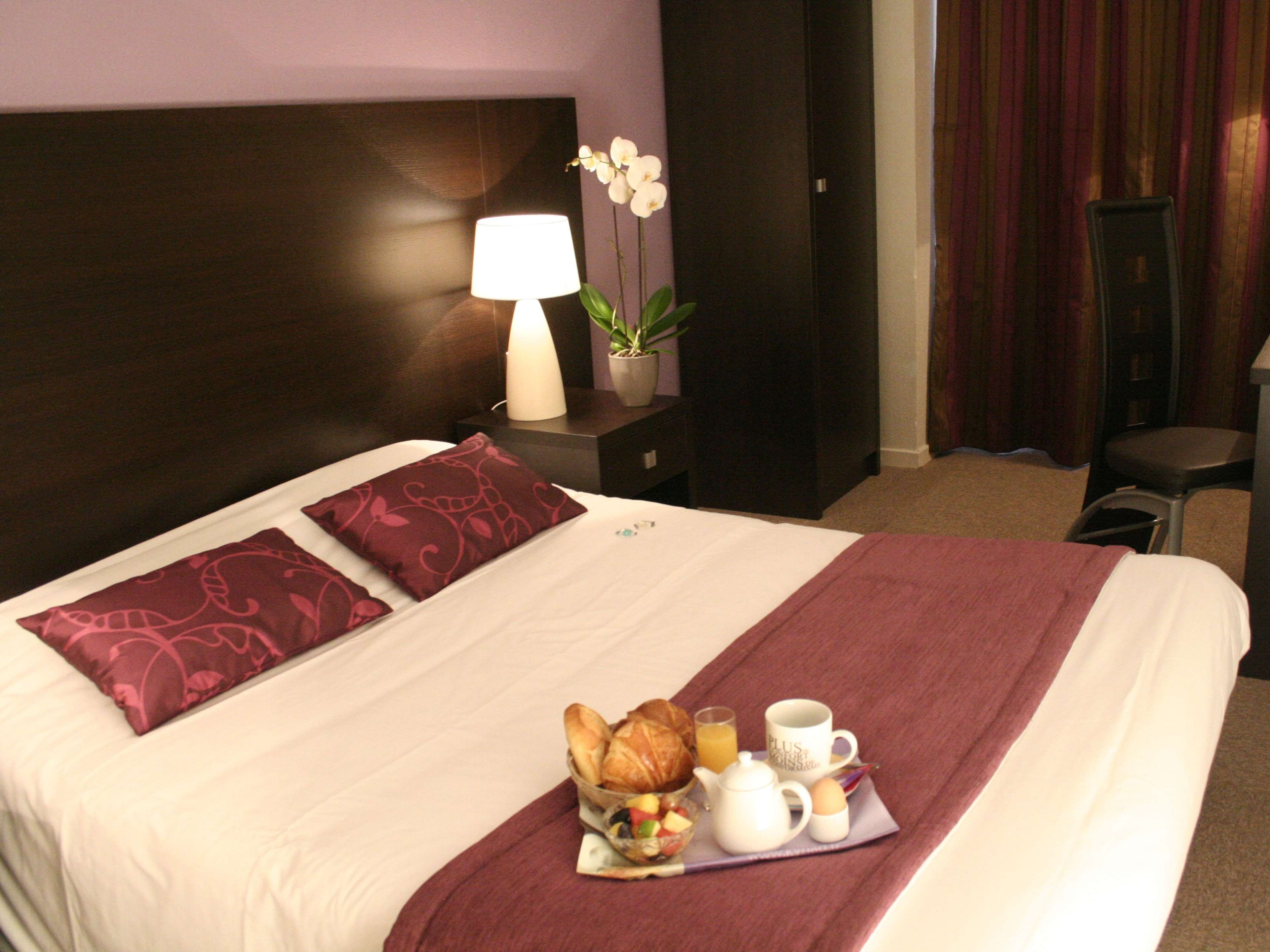 Una habitación de hotel con una cama, una mesita de noche con lámpara, una planta en maceta y una bandeja de desayuno sobre la cama.