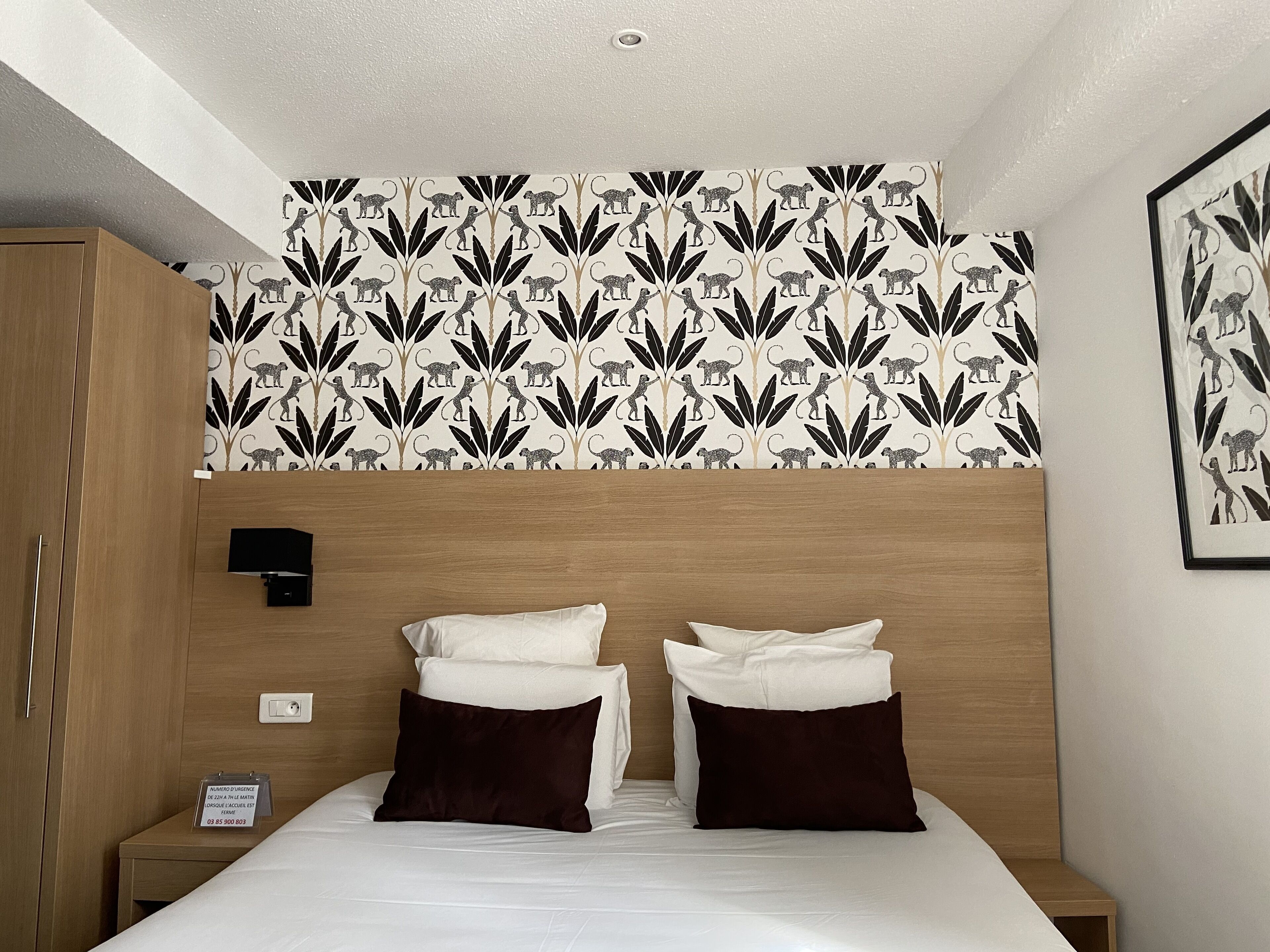 A hotel room with a bed, two pillows, a nightstand, a wall-mounted lamp, and a framed picture.