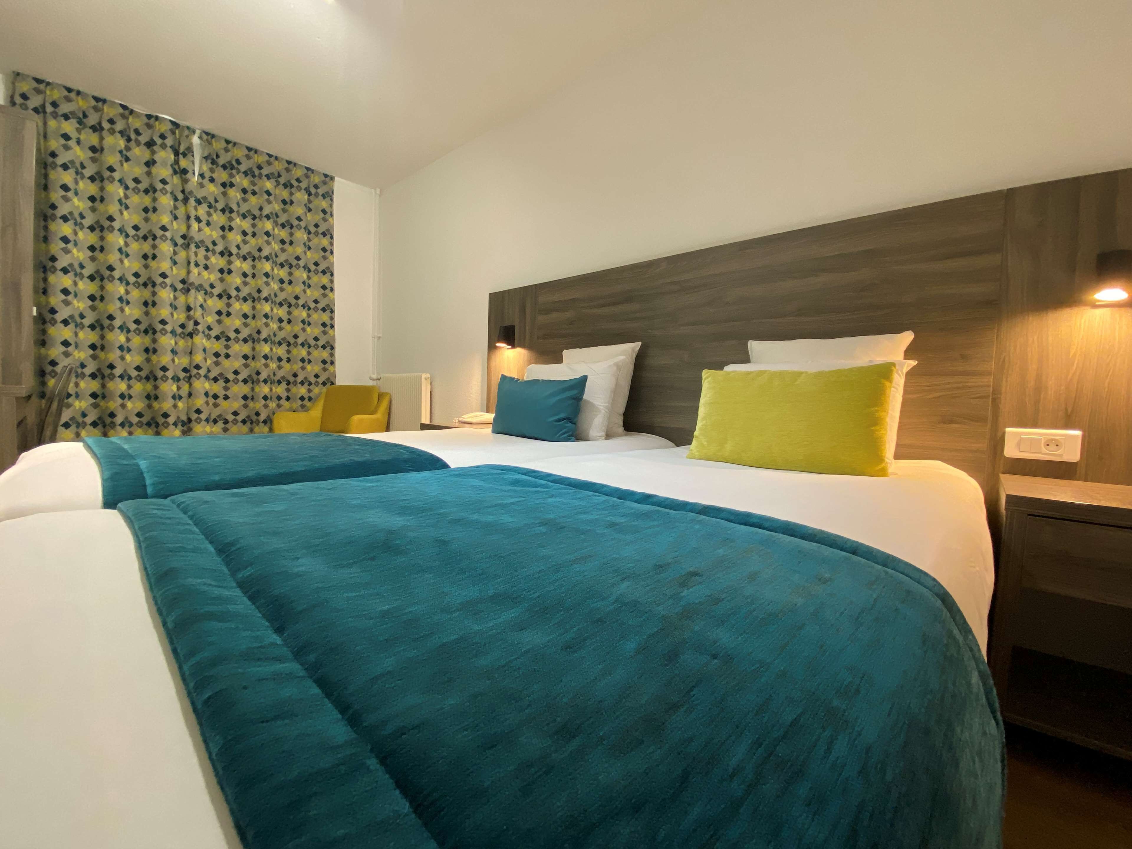 Habitación de hotel con dos camas, cabecero de madera, una silla amarilla y cortina estampada.