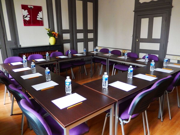 Sala de conferência com mesas compridas, cadeiras roxas, garrafas de água e papéis brancos.