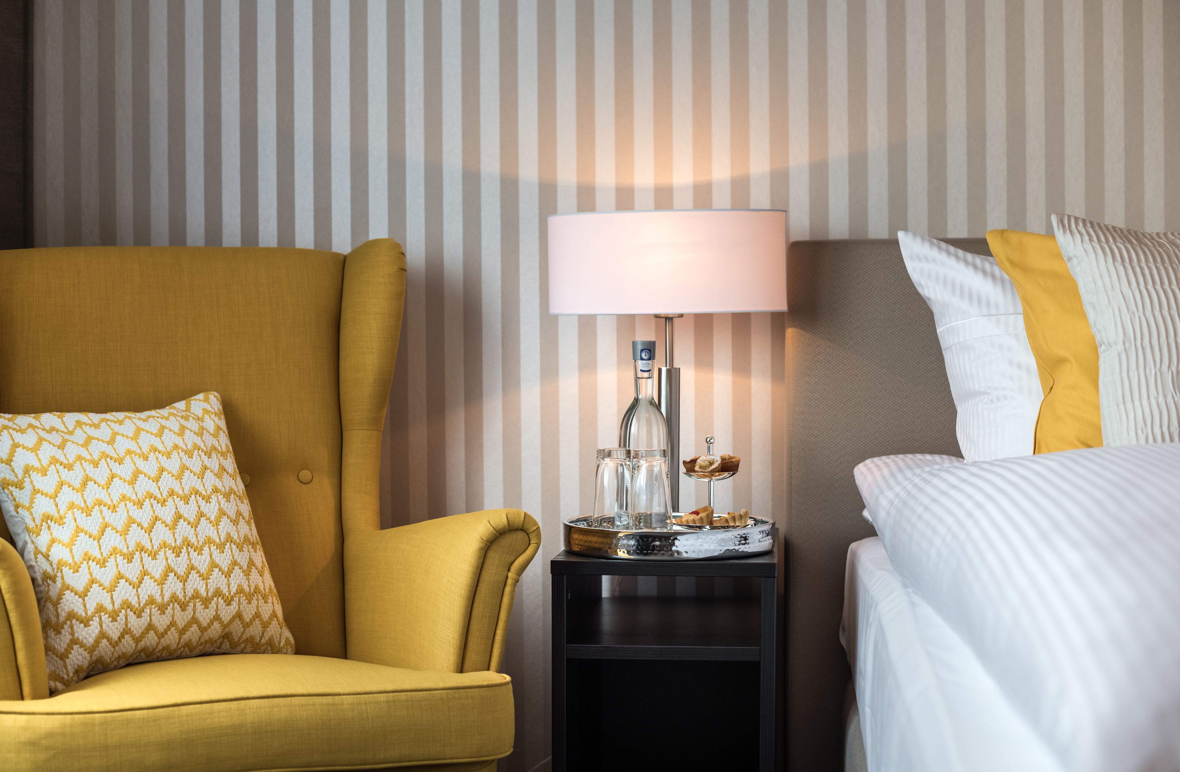 A hotel room with a yellow armchair, a bedside table with a lamp, and a bed with white bedding.