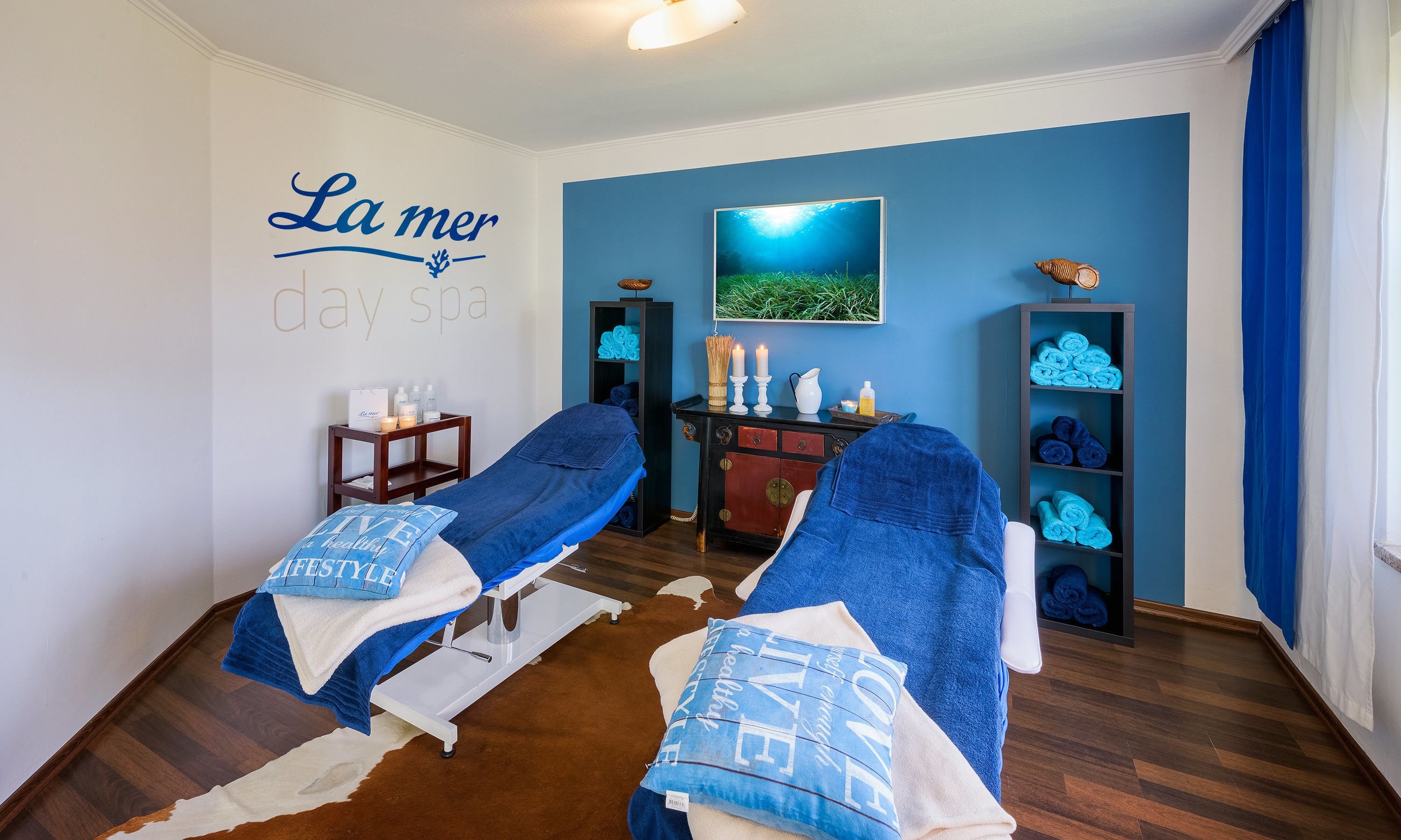 A spa room with two massage tables, a wall-mounted TV, and a blue wall with 'La mer day spa' signage.
