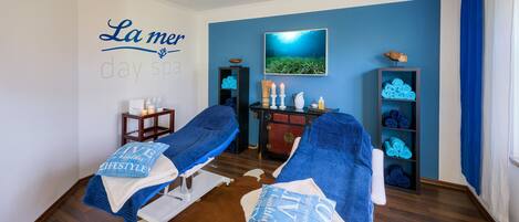 Une salle de spa équipée de deux tables de massage, d’un téléviseur fixé au mur et d’un mur bleu orné de l’enseigne « La mer day spa ».