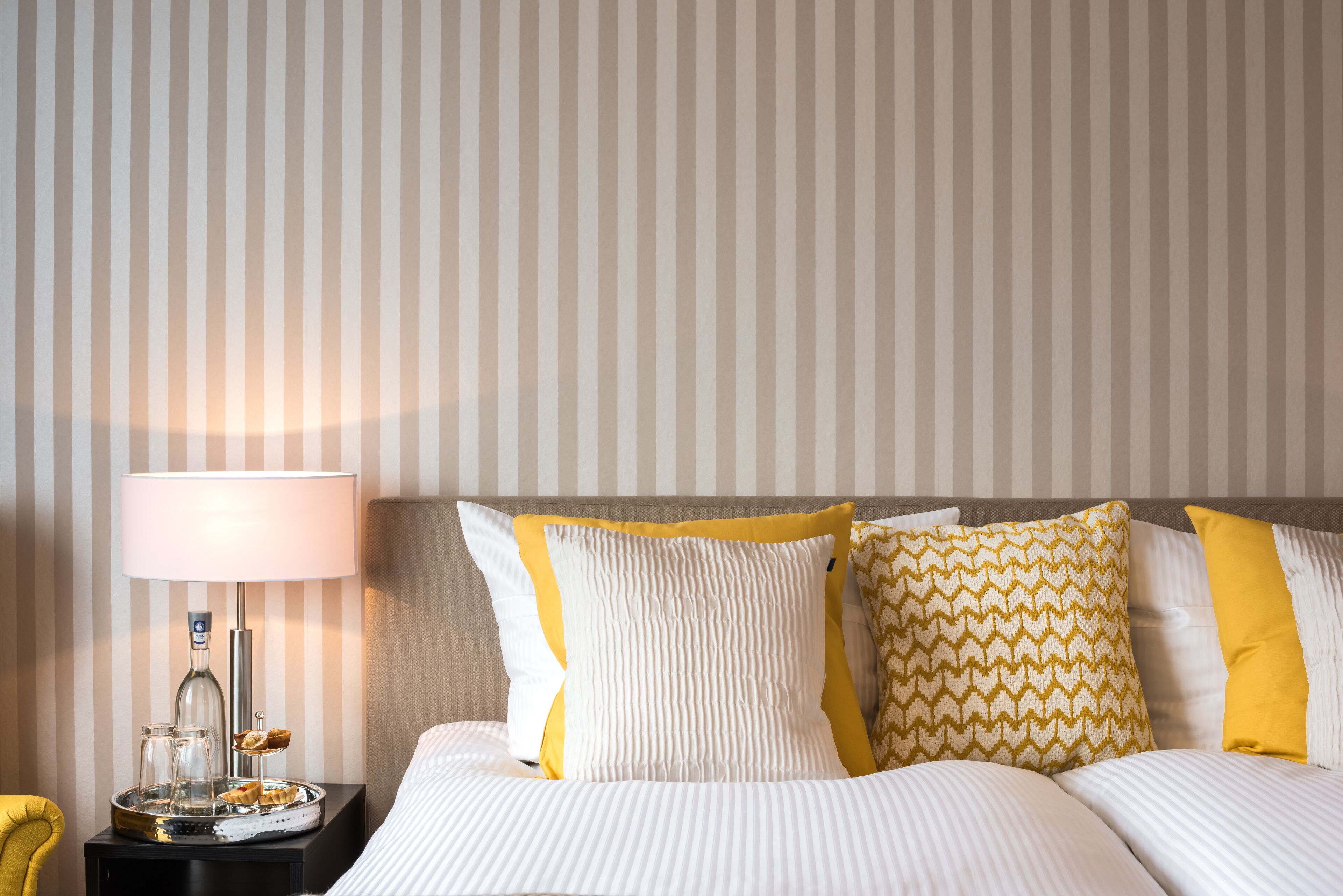 Een net opgemaakt bed met gestreept beddengoed, een bedlampje en een dienblad met glazen en een klein bord.