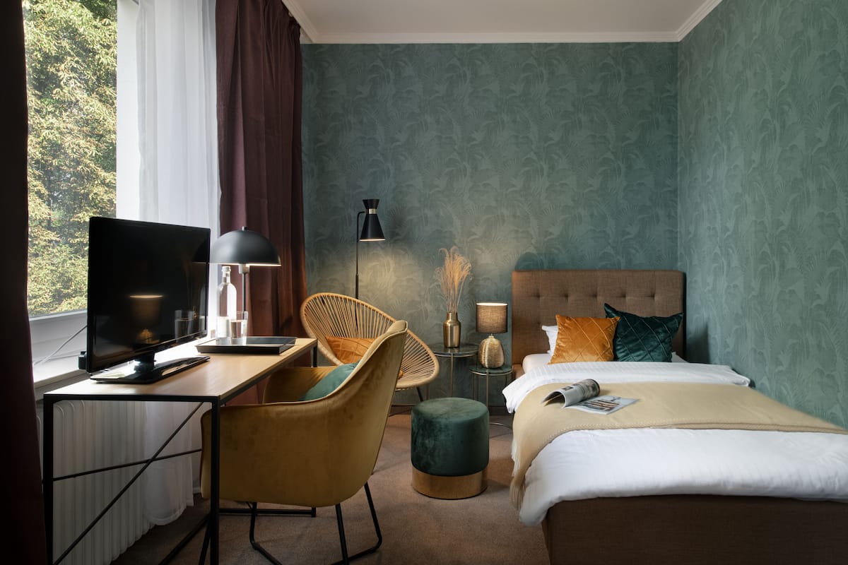 A hotel room with a bed, a desk with a computer, a chair, a lamp, and a window with curtains.