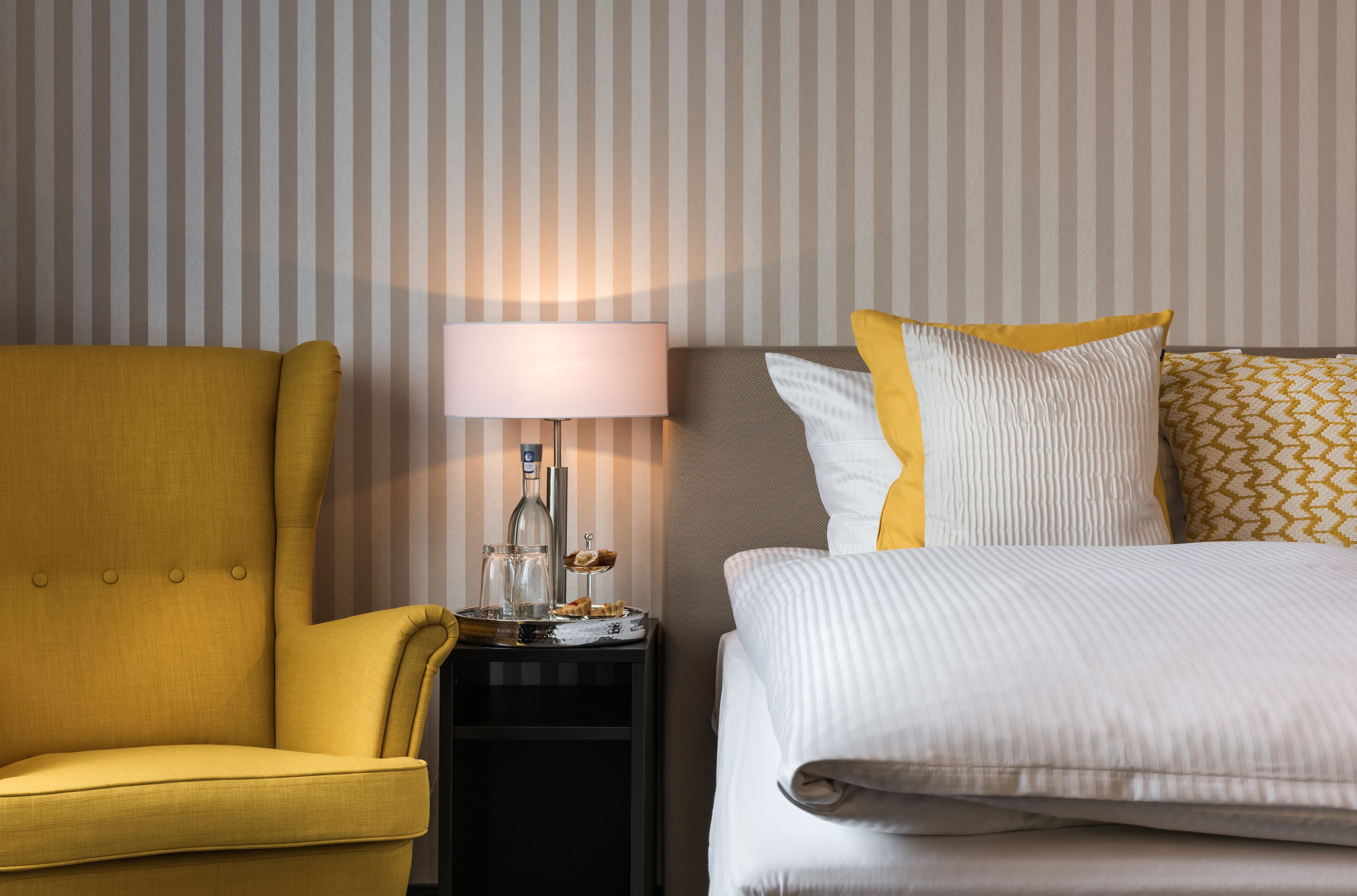 Una habitación de hotel con un sillón amarillo, una mesita de noche con lámpara y una cama con ropa de cama blanca y almohadas estampadas.