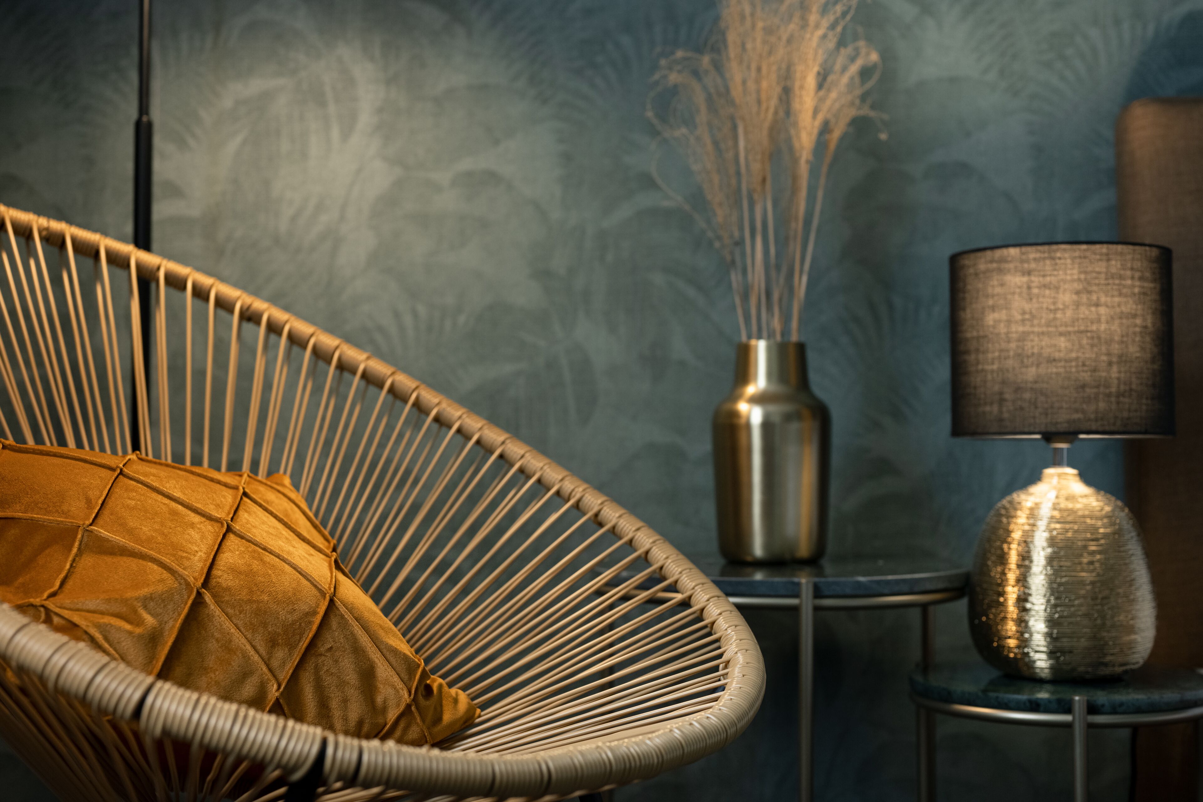 A wicker chair with a cushion, a vase with a plant, and a lamp on a side table.