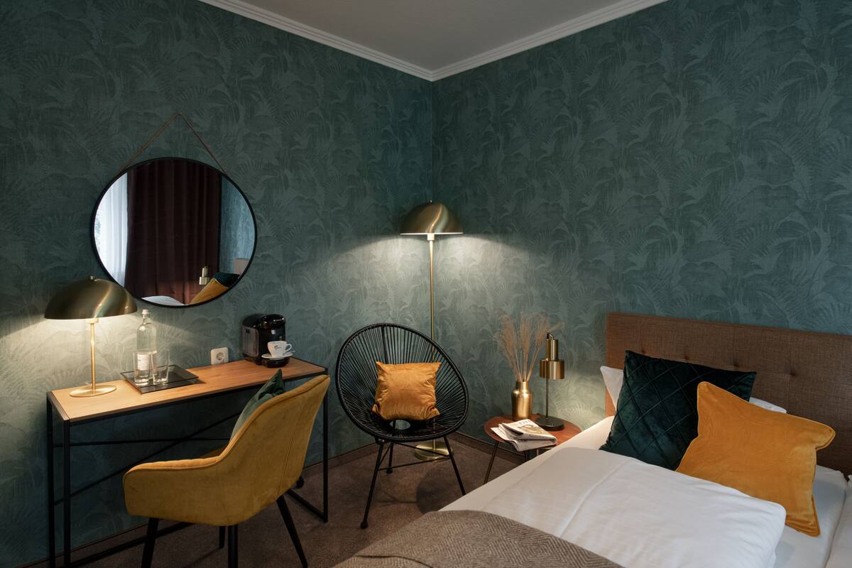 A hotel room with a bed, a desk, a chair, a round mirror, and a floor lamp.