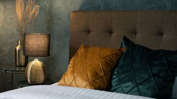 Cama bem arrumada com cabeceira estofada, abajur com cúpula texturizada e plantas secas decorativas.