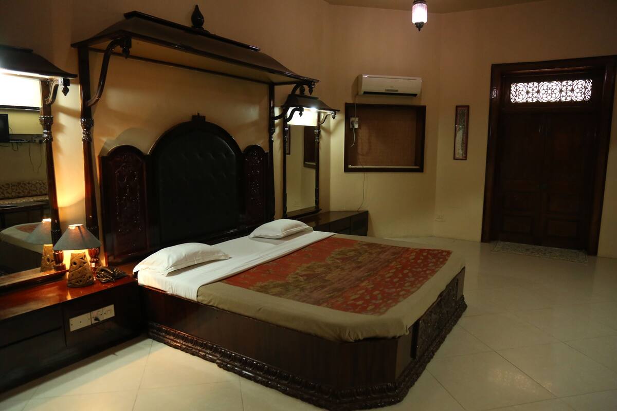 A traditional bedroom with a four-poster bed, a nightstand with a lamp, a dresser, and a large mirror.