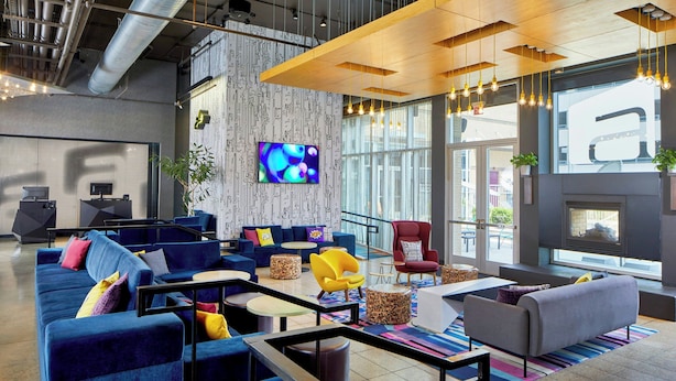 A modern lounge area with blue sofas, a red armchair, and a yellow chair. A flat-screen TV is mounted on the wall, and there are large windows providing natural light.