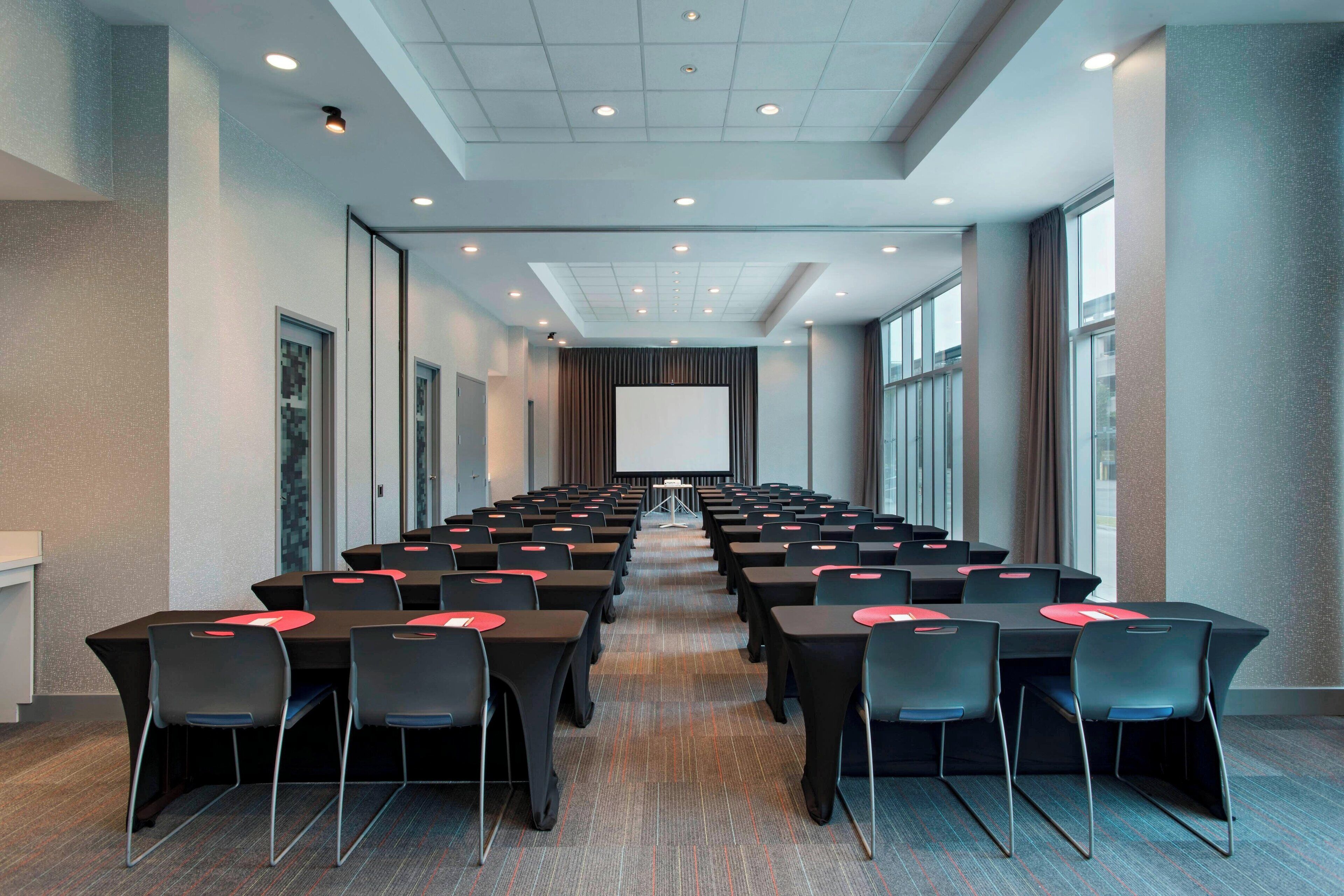 A conference room with rows of tables and chairs, a large screen at the front, and a carpeted floor.