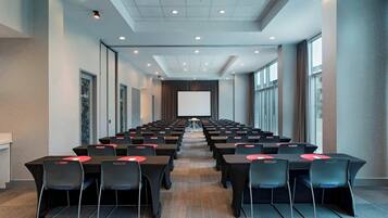 A conference room with rows of tables and chairs, a large screen at the front, and a carpeted floor.