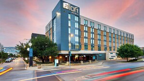 A modern hotel building with a prominent 'aloft' sign, large windows, and a visible outdoor seating area.