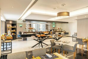 A modern hotel dining area with a central island, seating arrangements, and a kitchen in the background.
