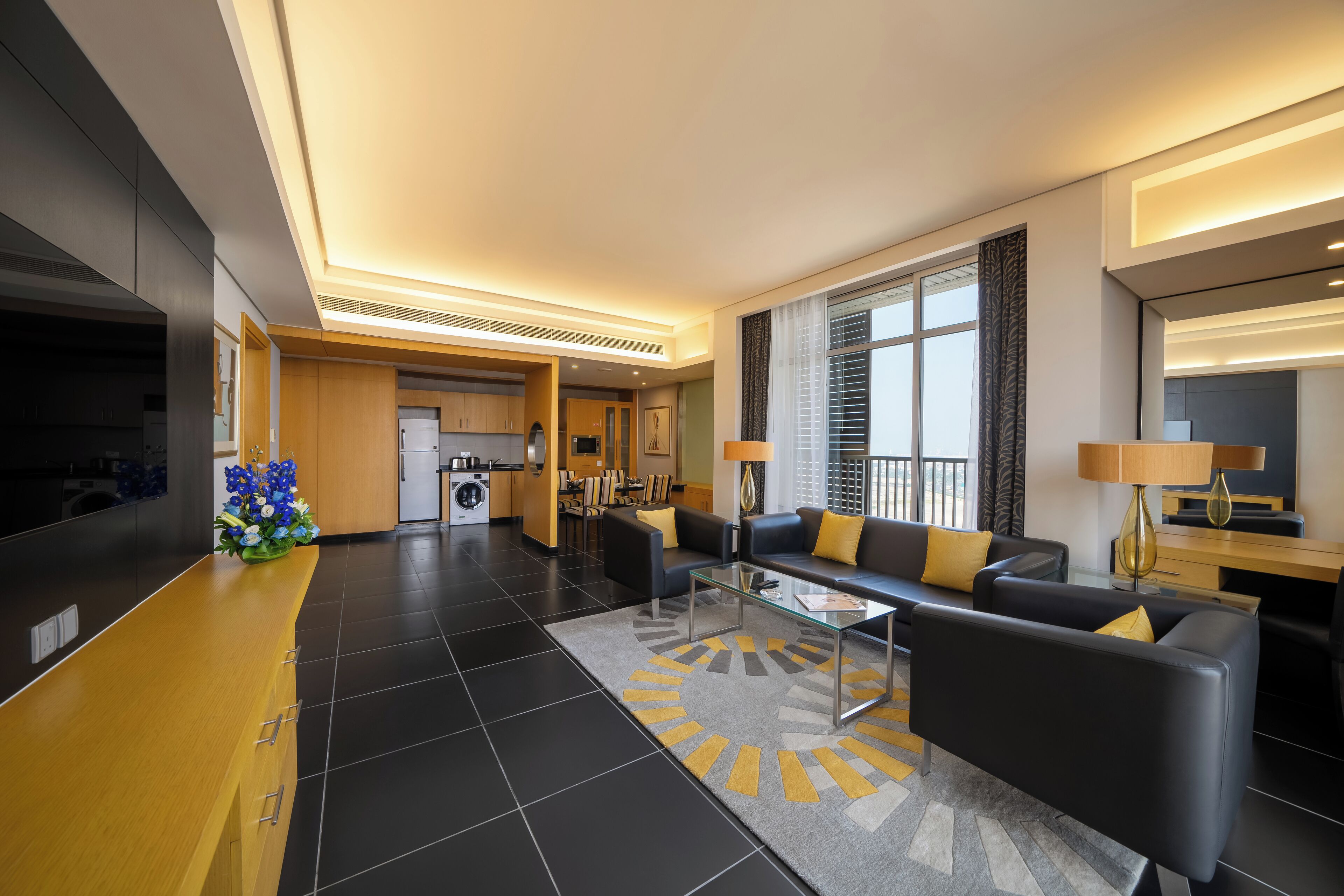 A modern hotel lobby with a central seating area, a kitchen in the background, and a large window providing natural light.