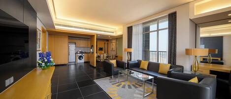 A modern hotel lobby with a central seating area, a kitchen in the background, and a large window providing natural light.