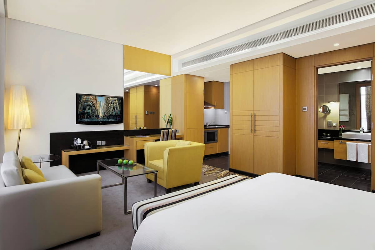 A hotel room with a bed, a sofa, a yellow armchair, a glass coffee table, a TV, and a kitchenette.