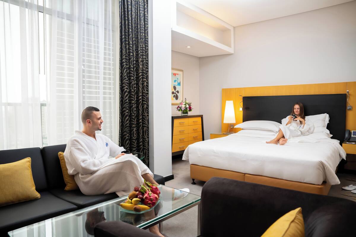 A hotel room with a bed, a sofa, a glass coffee table with fruit, and a nightstand with a lamp.