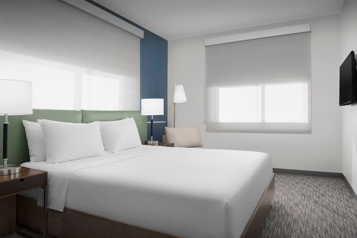 A hotel room with a large bed, two bedside tables, a flat-screen TV, and a window with blinds.