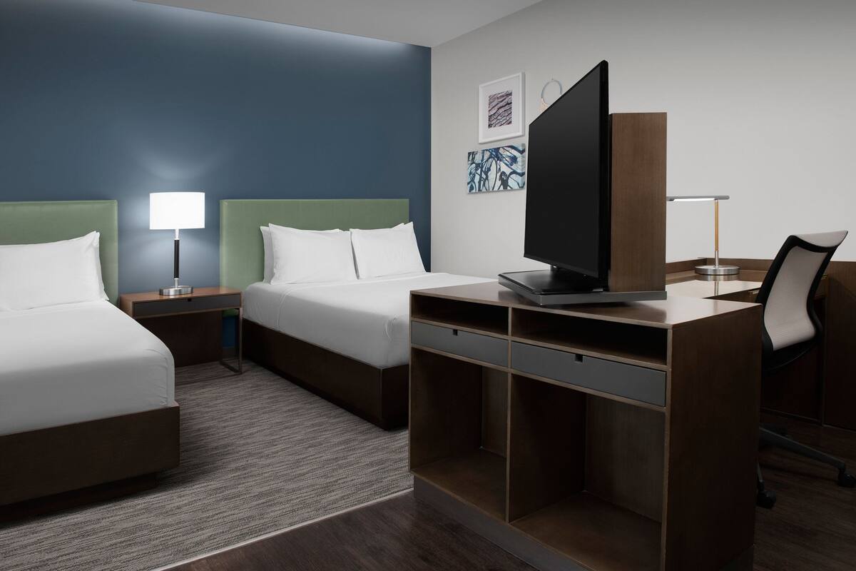 A hotel room with two beds, a desk, a flat-screen TV, and artwork on the wall.