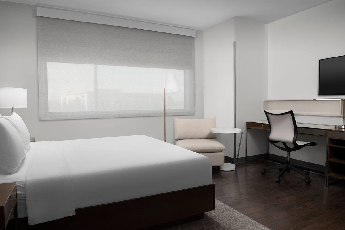 A modern hotel room with a large bed, a desk, a chair, a television, and a window with blinds.