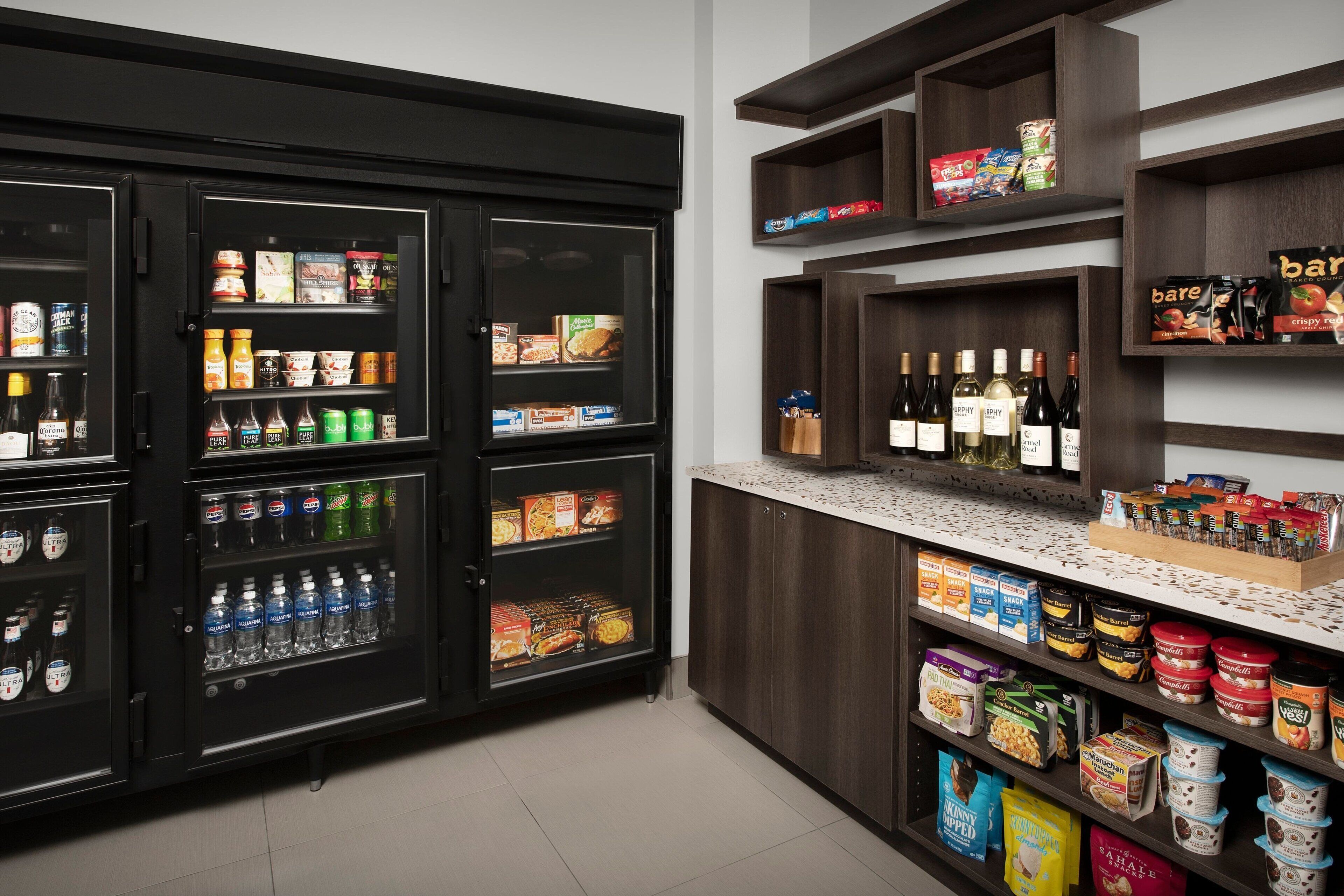 Une petite cuisine équipée d’un réfrigérateur rempli de boissons et d’en-cas, d’une cave à vin et d’étagères contenant des aliments emballés.