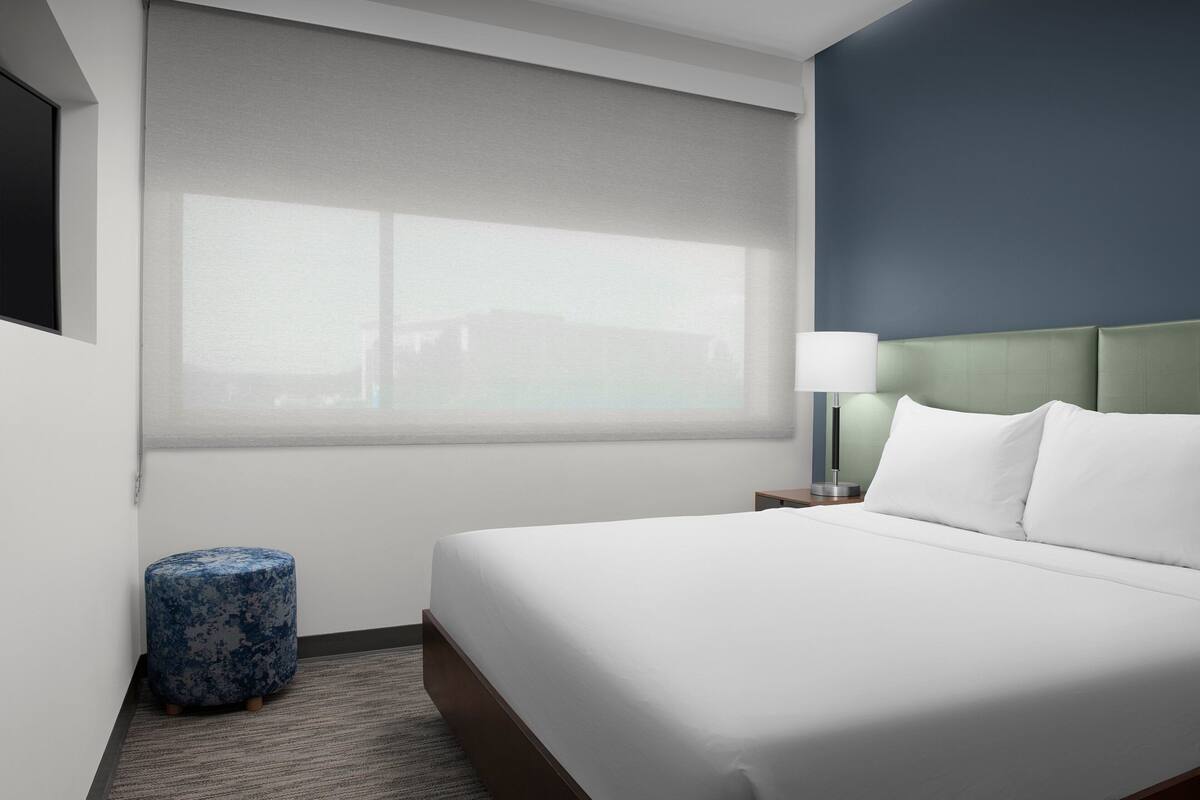 A hotel room with a bed, a nightstand, a lamp, a window with a view, and a blue ottoman.