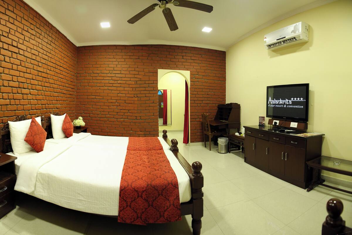 A hotel room with a brick wall, a bed with red and white bedding, a television, a desk with a computer, and a ceiling fan.