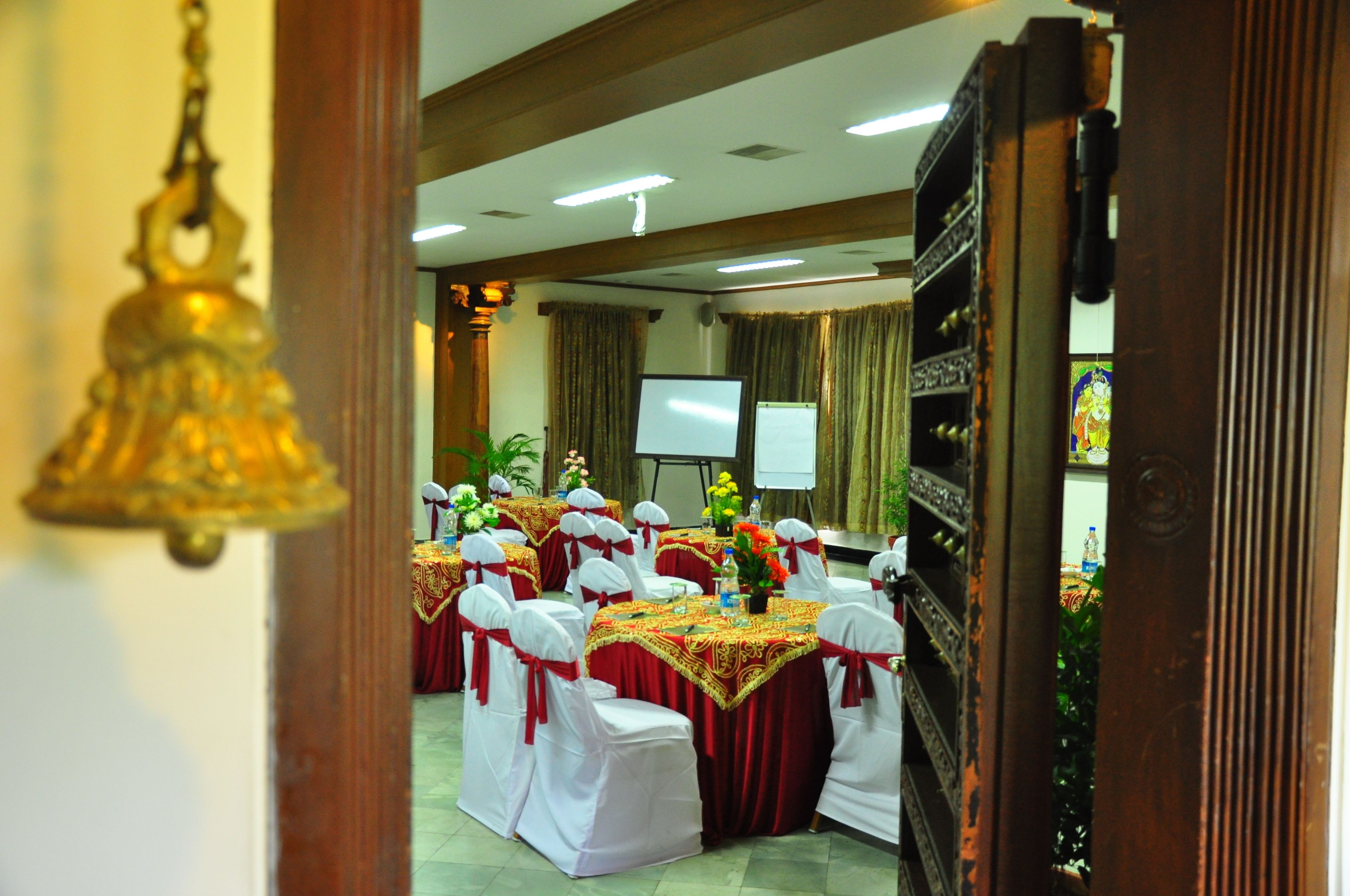 A banquet hall with tables set for a formal event, featuring white chairs with red sashes, a projector screen, and a floral arrangement.
