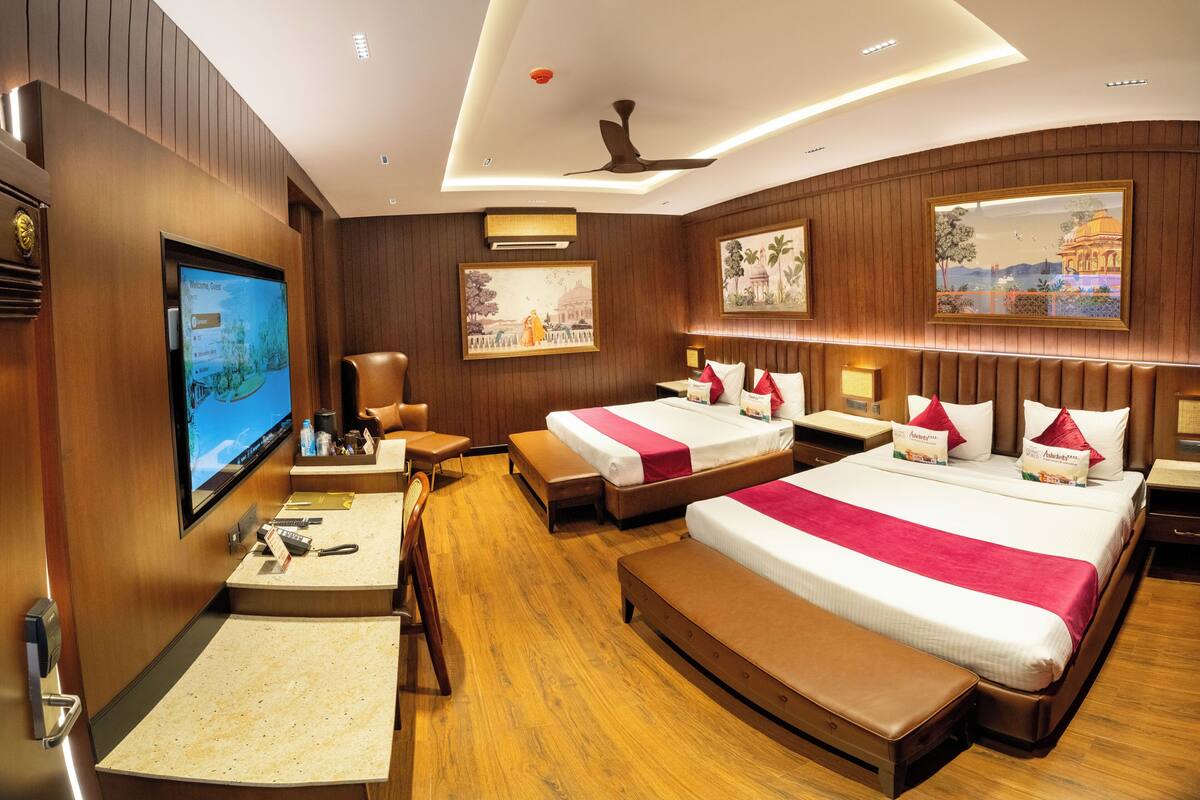 A hotel room with a TV, two beds, a desk, a chair, a bench, and a ceiling fan.