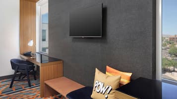 A hotel room with a flat-screen TV mounted on the wall, a desk with a chair, a bench, and a large window offering a city view.